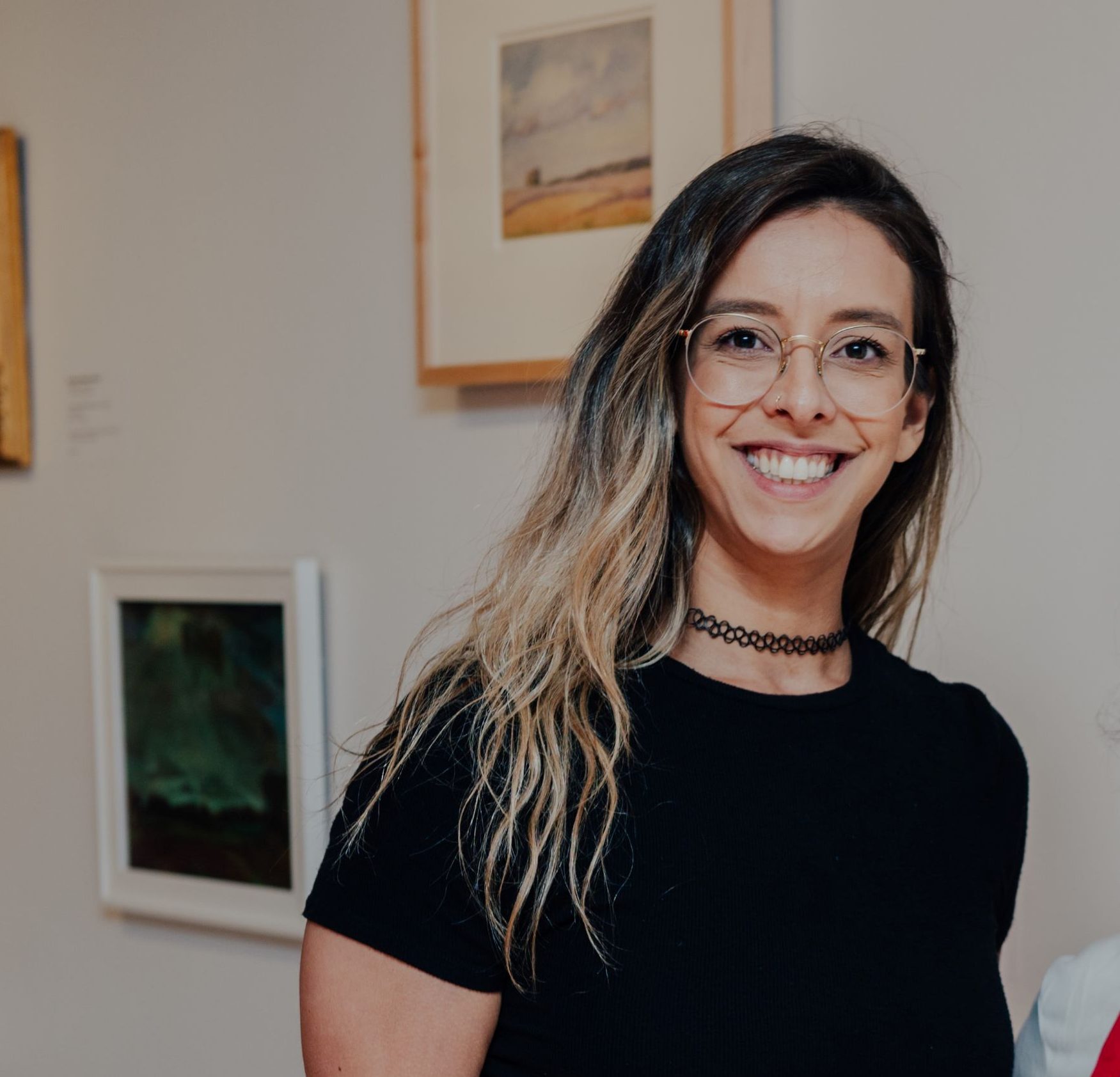 A woman with long hair smiling. She is wearing a black t-shirt, a black necklace, and gold glasses.