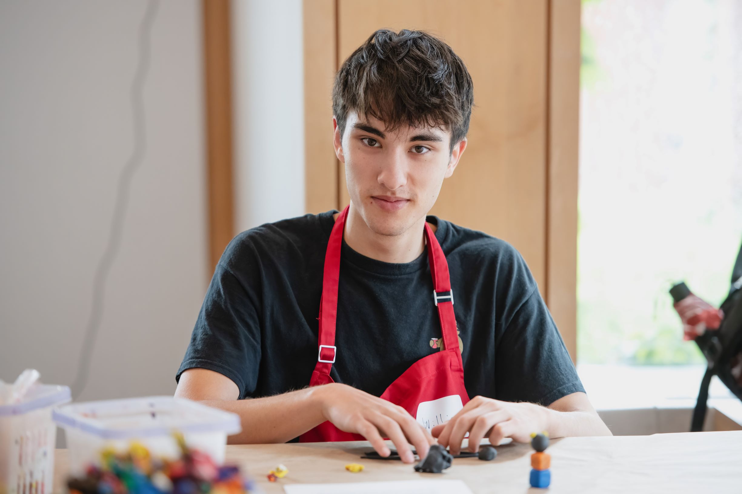 A young man in a red apron is working with clay at a table.