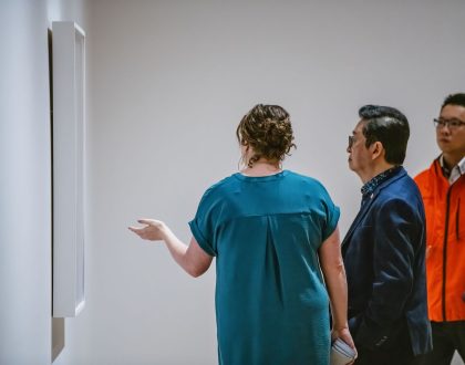 A woman in a blue dress is giving a tour to two men in front of an artwork in a gallery.