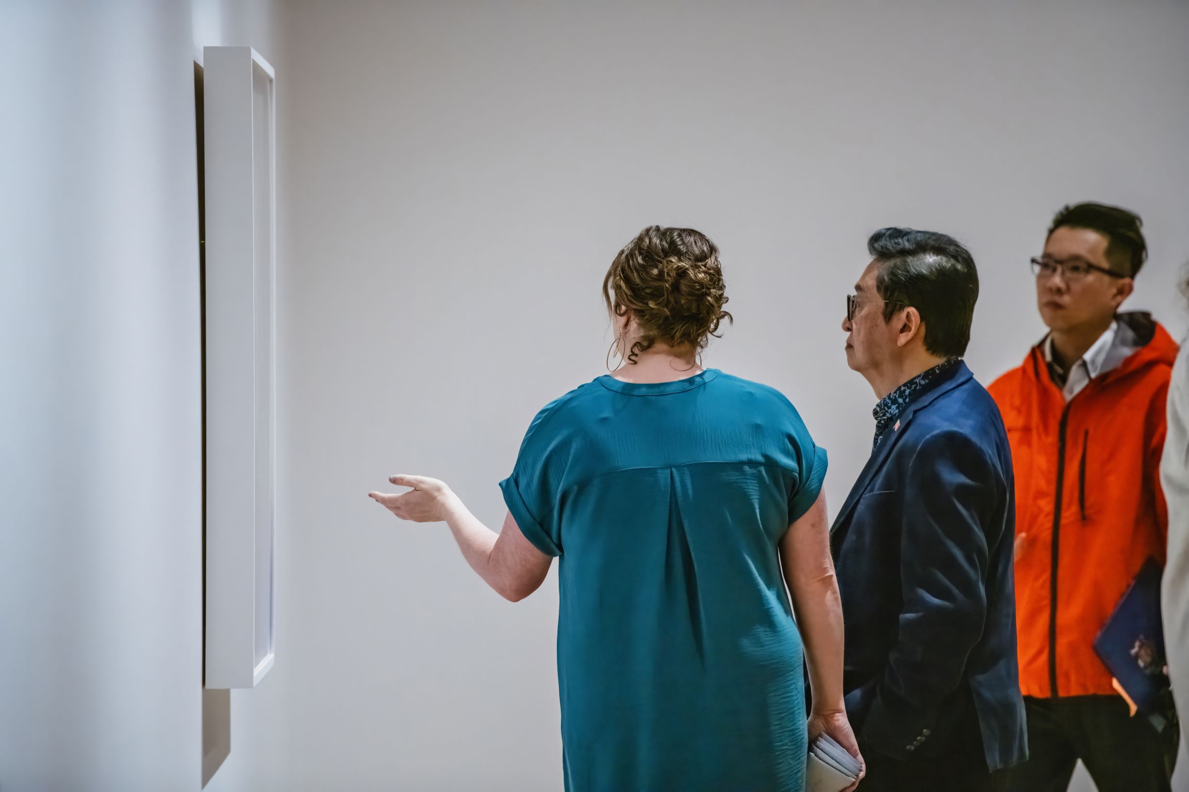 A woman in a blue dress is giving a tour to two men in front of an artwork in a gallery.