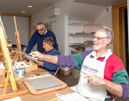 A man with silver beard and hair is smiling while painting on a canvas in a paint studio. Behind him, a woman and a man are looking at a paper and discussing.