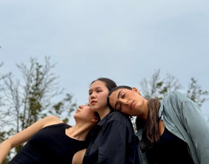 Three female dancers are leaning towards each other with their upper bodies, set against a backdrop of trees.