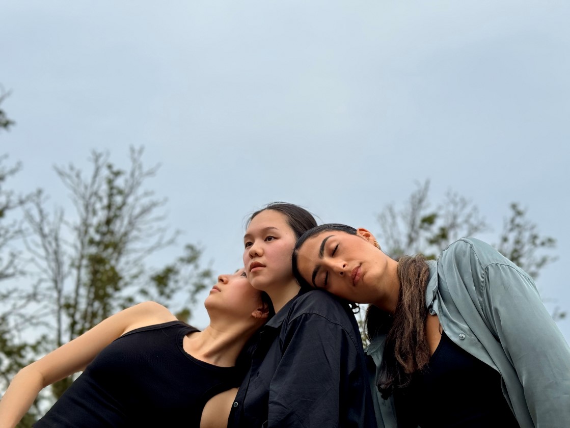 Three female dancers are leaning towards each other with their upper bodies, set against a backdrop of trees.