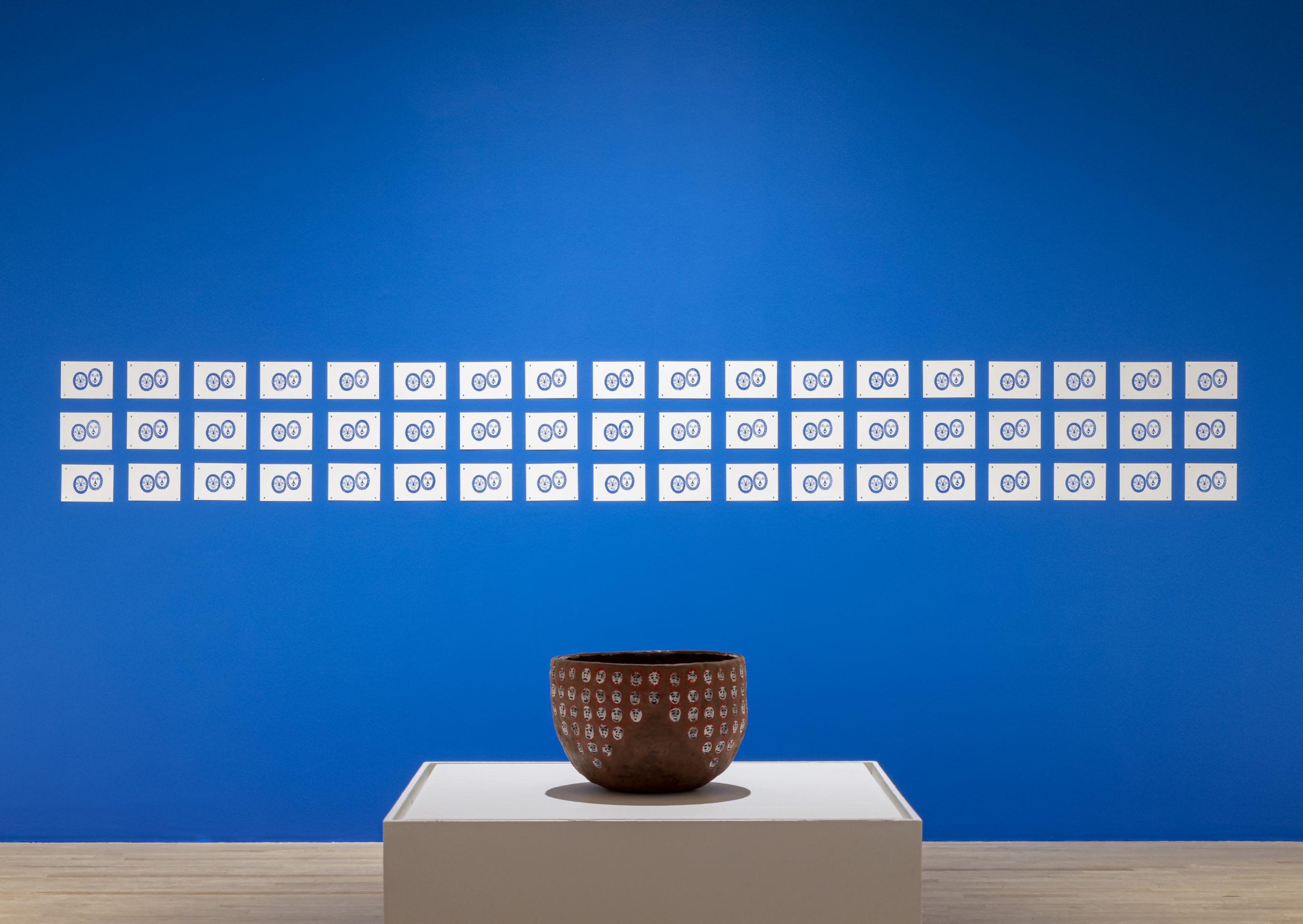 In an exhibition space. Background is a striking blue wall with repeating printmaking artwork on. A ceramic artwork placed at the front.