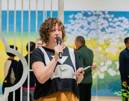 A woman is holding a mic and giving a speech, in a gallery setting. There is Chinese landscape painting and visitors viewing the art in the background.