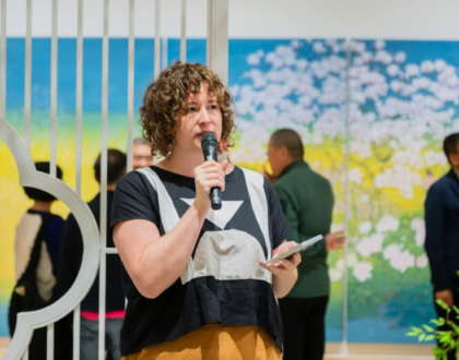 A woman is holding a mic and giving a speech, in a gallery setting. There is Chinese landscape painting and visitors viewing the art in the background.