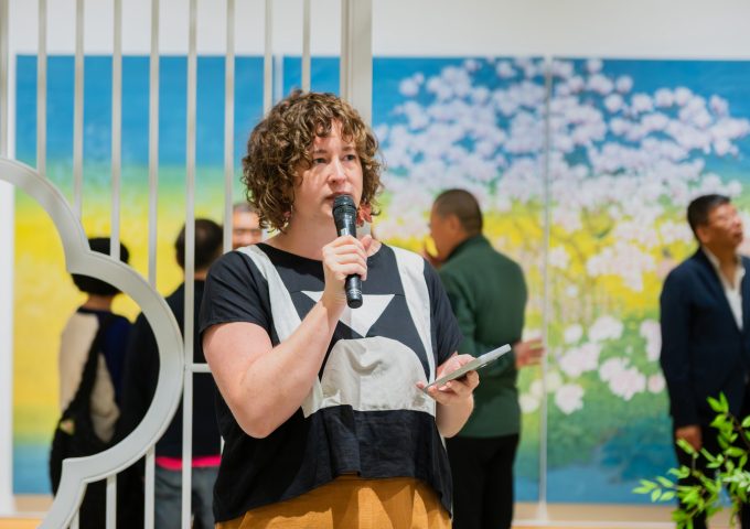 A woman is holding a mic and giving a speech, in a gallery setting. There is Chinese landscape painting and visitors viewing the art in the background.
