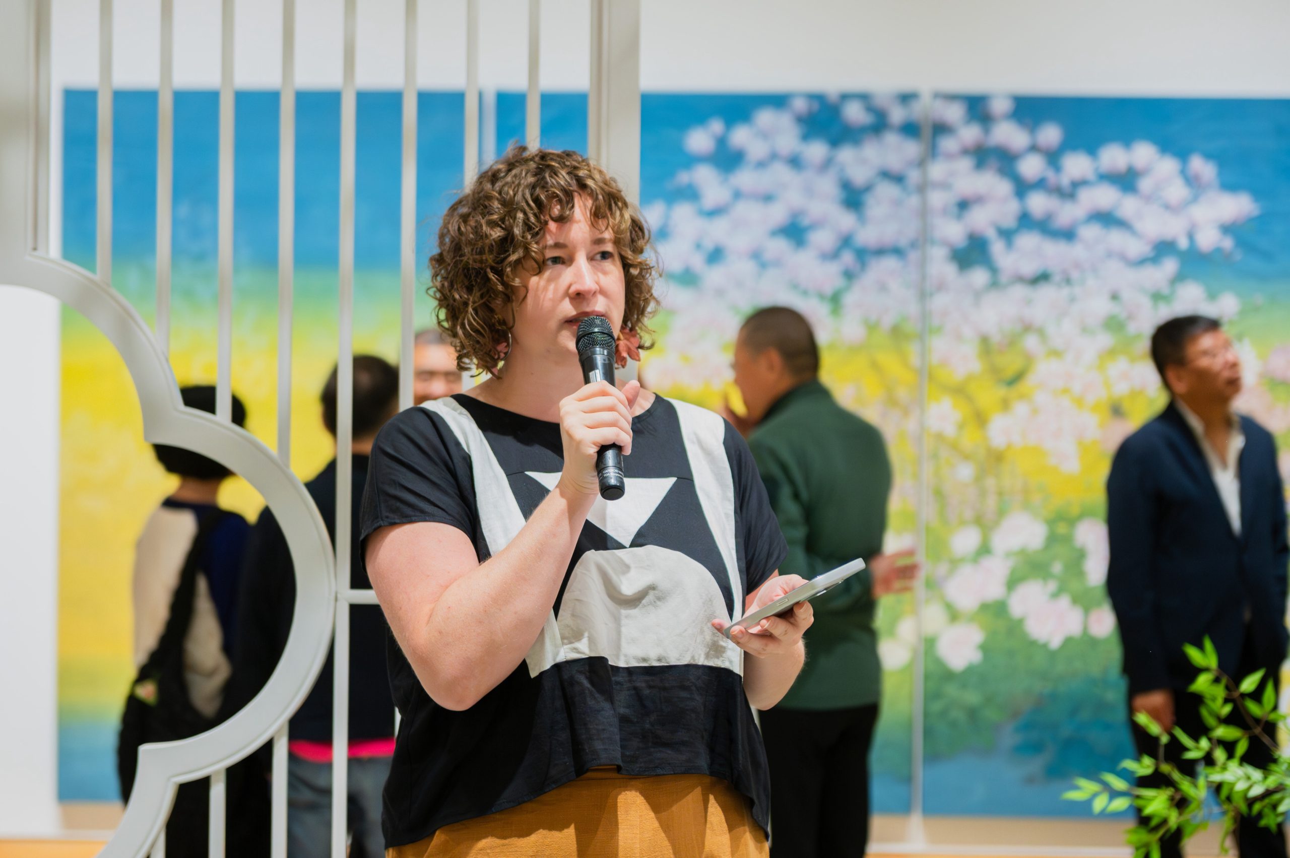A woman is holding a mic and giving a speech, in a gallery setting. There is Chinese landscape painting and visitors viewing the art in the background.
